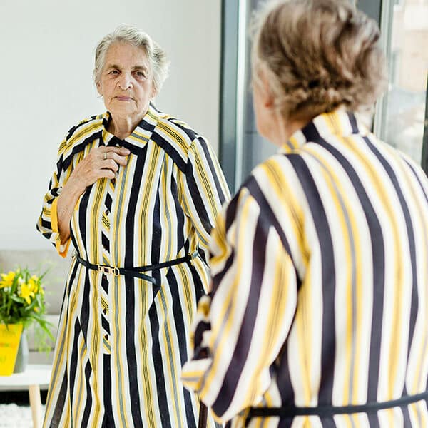 Das Bild zeigt eine ältere Frau vor einem Spiegel, welche ihre Kleidung zurecht rückt.